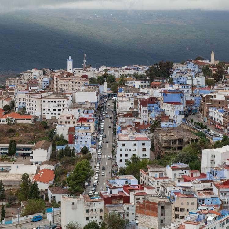 An elevated, high-angle view of the blue-washed buildings and streets of Chefchaouen nestled below mountains in northern Morocco.