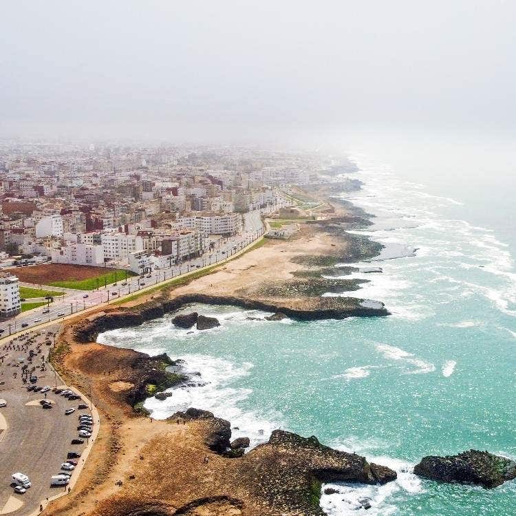 An aerial view of the rugged, rocky coastline and city buildings along the misty Atlantic Ocean corniche in Rabat, Morocco.