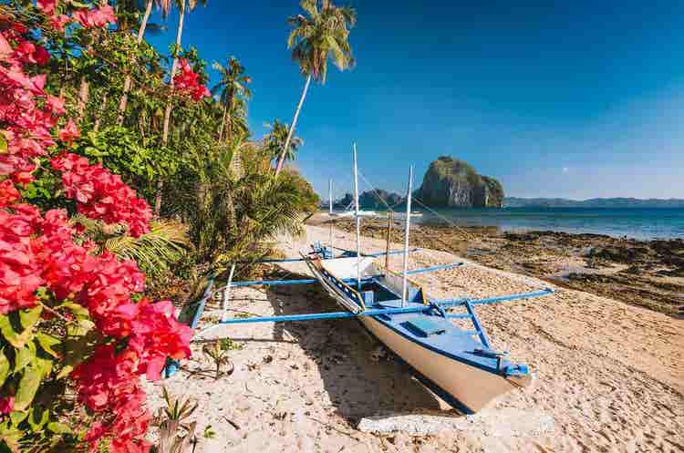 Traditional wooden boat resting along pristine tropical beach with turquoise waters, soft golden sand, clear skies, and peaceful coastal scenery.