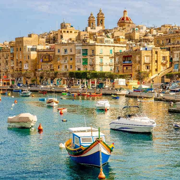 Colorful traditional Maltese luzzu boats float in the calm blue harbor, backed by sunlit limestone buildings and a domed church.
