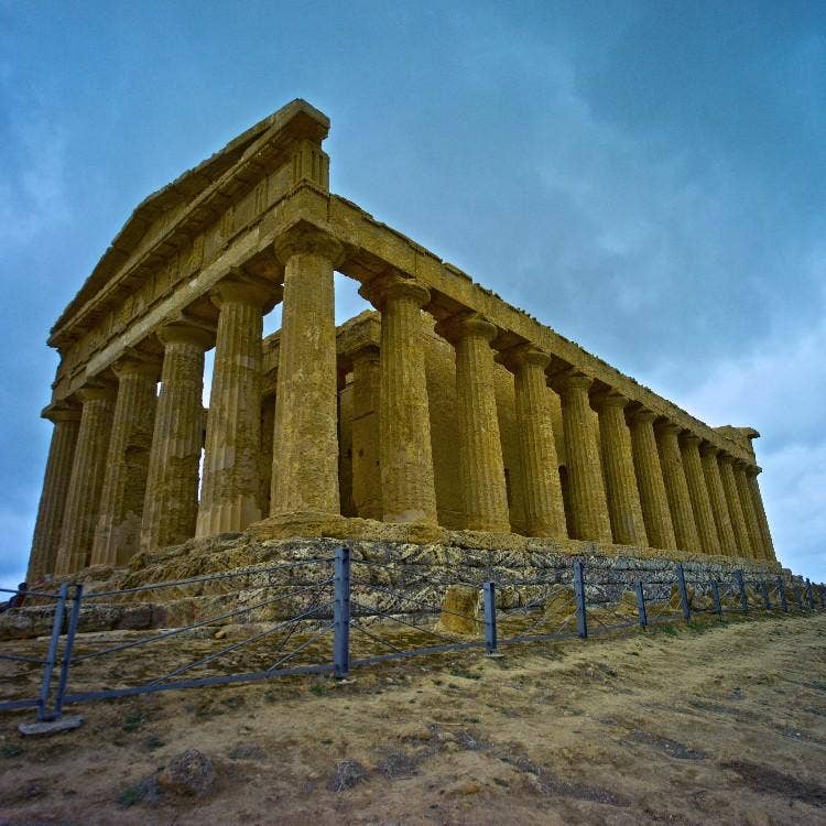 A low-angle shot of the ancient Greek Temple of Concordia in Agrigento, Sicily, highlighting its standing Doric columns under a cloudy sky.