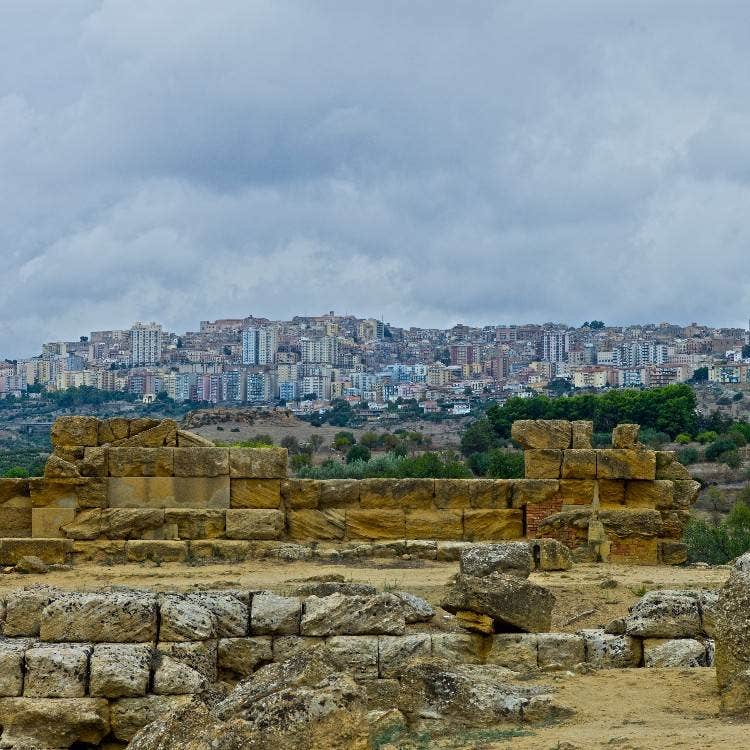 Ancient stone ruins in the foreground with the modern city of Agrigento built on a hillside under a cloudy sky.