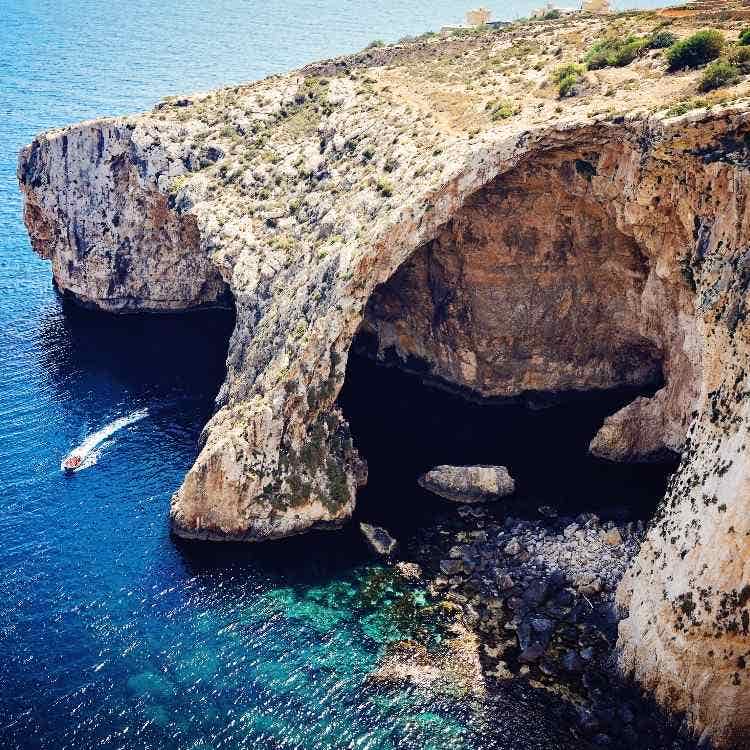 A high-angle view of the massive natural sea arch at the Blue Grotto in Malta, featuring turquoise water and a small boat.
