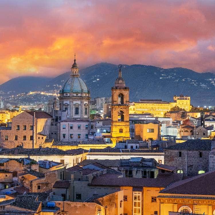 A panoramic evening view of Palermo's historic skyline under a dramatic sunset, showcasing illuminated domes, towers, and mountain backdrops.
