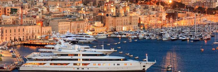 A panoramic evening view of Malta's historic Grand Harbour, showcasing luxury yachts docked alongside illuminated traditional Maltese limestone waterfront buildings.
