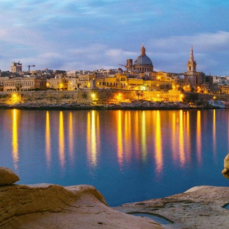An evening view of Valletta's illuminated skyline reflecting in Marsamxett Harbour, featuring the iconic dome of Mount Carmel Basilica.