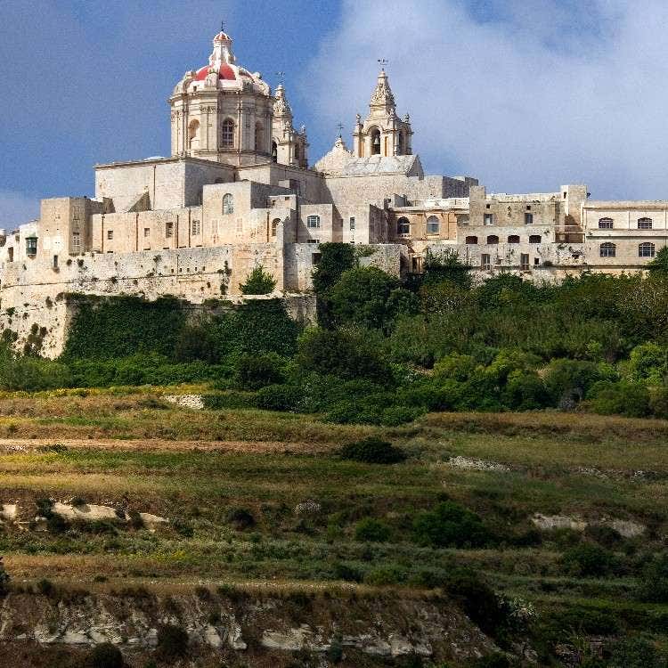 A distant hillside view of the fortified medieval city of Mdina, Malta, highlighting the prominent dome of St. Paul's Cathedral.