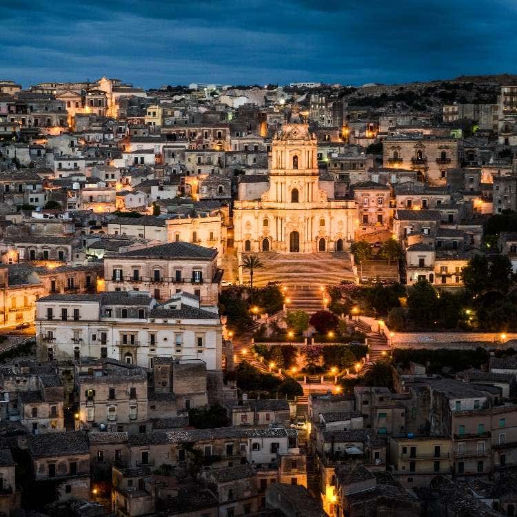 A panoramic night view of the illuminated Baroque cathedral of San Giorgio rising above the historic town of Modica, Sicily.