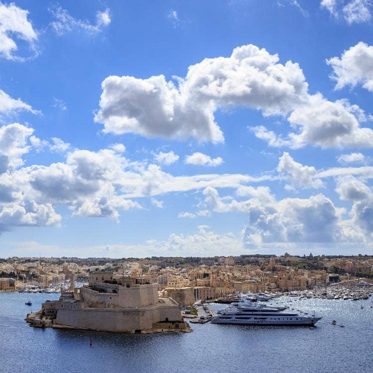 A panoramic view of Malta's historic Grand Harbour under a cloudy sky, showcasing fortified stone bastions and luxury yachts docked nearby.