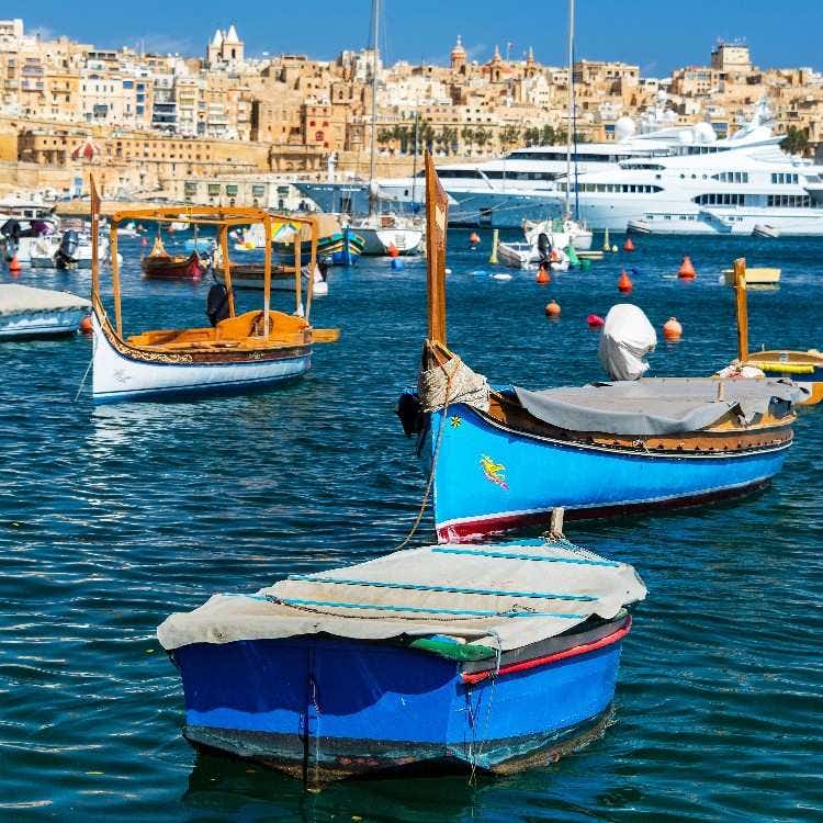 Traditional wooden dgħajsa boats bobbing in the blue water of Malta's Grand Harbour, with historic limestone buildings behind them.