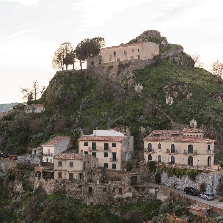 The medieval hillside village of Savoca, Sicily, featuring stone buildings and historical structures terraced on a steep green hill.