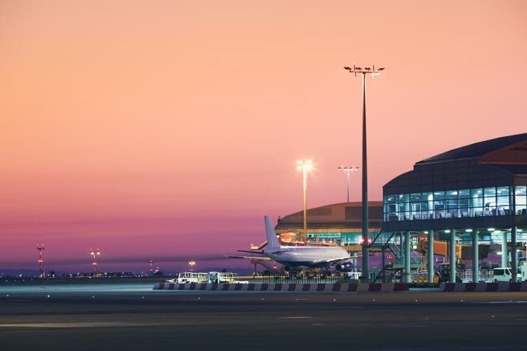 An airplane parked at a modern airport terminal gate at dawn, with a vibrant pink and orange sky overhead.